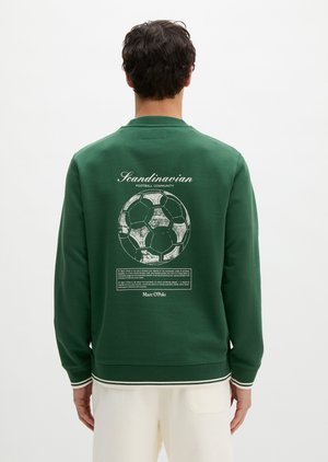 Person som har på seg grønn genser med hvit grafikk av en fotball og teksten "Scandinavian Football Community" på baksiden.