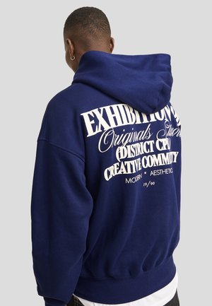 Navy blue hoodie featuring large white text graphic on back, textured fabric, adjustable hood, and ribbed cuffs and hem.