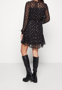 Eine Person in einem schwarzen, gemusterten Kleid mit Rüschen und langen Ärmeln, die kniehohe schwarze Stiefel trägt, steht vor einem weißen Hintergrund.