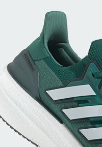 Tmavě zelená sportovní bota s texturovaným síťovaným svrškem, třemi bílými pruhy a bílou polstrovanou podrážkou, na které je označení "ULTRABOOST."