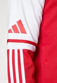 Rood en wit sportshirt met een gestructureerde stof, drie witte strepen op de mouw en een opvallend rood Adidas-logo.