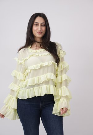 Frau mit langen dunklen Haaren, die eine blassgelbe durchsichtige Bluse mit mehreren Rüschenlagen und dunkelblaue Jeans trägt, steht vor einem hellen Hintergrund.