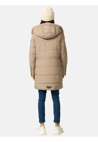 Beige pufferjas met een capuchon, gewatteerd ontwerp en logo-detail aan de onderrand, gecombineerd met blue jeans en witte sneakers.
