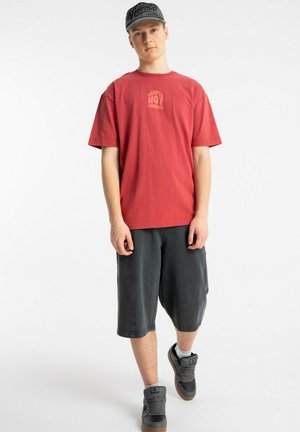 Młody mężczyzna ubrany w czerwoną koszulkę z napisem "Waves Not Worries", ciemne krótkie spodenki do kolan, szare tenisówki i szarą czapkę z daszkiem, stojący na białym tle.