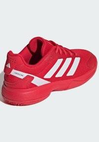 Röd tennissko med ett strukturerat mesh-övre, vit tre-stripesdesign, vadderad krage och strukturerad gummisula för bättre grepp.