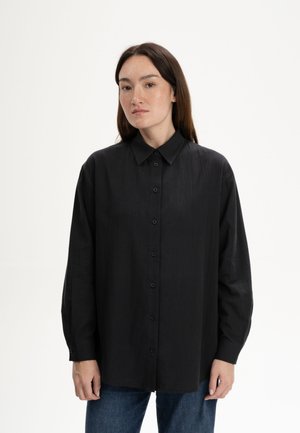 Frau mit langen dunklen Haaren, die ein übergroßes schwarzes Hemd mit Knöpfen und blaue Jeans trägt und vor einem schlichten weißen Hintergrund steht.