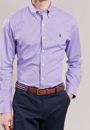 Mann trägt ein violettes kariertes Hemd mit Knöpfen, das in eine marineblaue Hose gesteckt ist, dazu einen braunen Ledergürtel und eine gestreifte Stoff-Gürtelschlaufe.