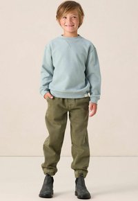 Blauwe fleece sweater, relaxte pasvorm, ribbelmanchetten. Olijfgroene cargo broek met elastische enkels. Zwarte enkellaarsjes. Eenvoudige achtergrond.
