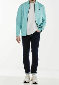 Light blue zip-up jacket with black trim, white crew neck shirt, dark denim jeans, and beige sneakers with white accents.