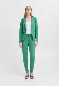 Groene blazer met bijpassende joggers, gemaakt van zachte stof. De blazer heeft een enkele knoopsluiting, de joggers hebben een elastische tailleband en een koord.