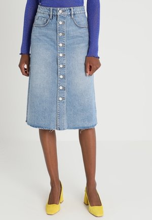 Kvinde iført en knap-front, knælængde denimnederdel, lilla langærmet top og gule sko med blokhæl, stående på en hvid baggrund.