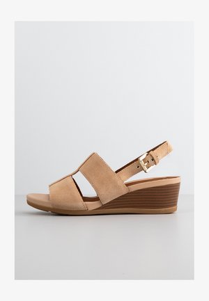 Beige ruskinds kilehæle sandaler med to brede overremme, justerbar ankelrem med guldfarvet hardware, og en formet brun sål med et tekstureret mønster.