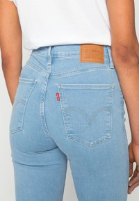 Ljusblå denimjeans med hög midja, med en läderlappar bak, två bakfickor och en röd etikett på sidan.