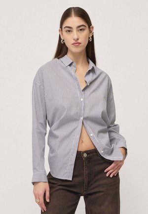 Donna con capelli castani lisci che indossa orecchini a cerchio in argento e una camicia sottile a righe, larga e parzialmente sbottonata, con pantaloni scuri a vita alta.
