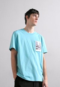 Svijetloplava pamučna majica s džepom na prsima i tiskom fotografije. Kratki rukavi i opušteni kroj. Glatka tekstura s minimalnim detaljima.