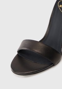 Μαύρη δερμάτινη ψηλοτάκουνη σандάλη με φαρδύ λουράκι στο δάχτυλο, ομαλή υφή και κομψά περιγράμματα. Η εσωτερική επένδυση είναι φτιαγμένη από σκούρο υλικό, με ελάχιστες λεπτομέρειες.