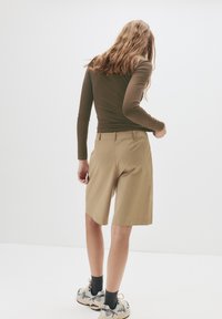 Khaki skræddersyede shorts med en afslappet pasform, parret med en tætsiddende brun langærmet T-shirt. Modellen har sporty sneakers og mørke strømper på.