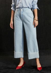Jeans de pierna ancha color azul claro con un dobladillo deshilachado y diseño de cinco bolsillos; combinados con tacones rojos de punta. Rayas verticales en la camisa.