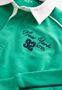 Groene poloshirt met een witte kraag, voorzien van geborduurd "New York 92 ATHL" in marineblauw. Zachte katoen met ribbelaccenten.