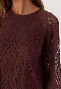 Bordeaux spetsblus med långa ärmar, som har intrikata blommönster och en texturerad yta. Rund halsringning, semi-transparent tyg.