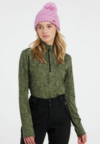 Majica dugih rukava u maslinasto zelenoj boji s crnim točkama, s polu-zip ovratnikom. Uparen s crnim hlačama i ružičastim pletenim šeširićem.