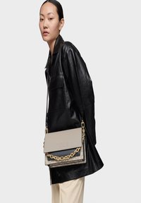 Mujer viste una chaqueta de cuero negra y sostiene un bolso TOUS beige y negro con una cadena dorada, contra un fondo claro y sencillo.