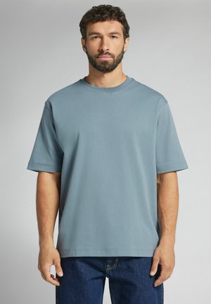 Mand med kort krøllet hår og skæg iført en ensfarvet lyseblå kortærmet skjorte og mørkeblå jeans, stående mod en grå baggrund.