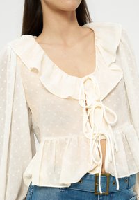 Woman wearing a cream, sheer, long-sleeve blouse with ruffled collar, tie-front detail, and light polka dot texture paired with blue jeans.