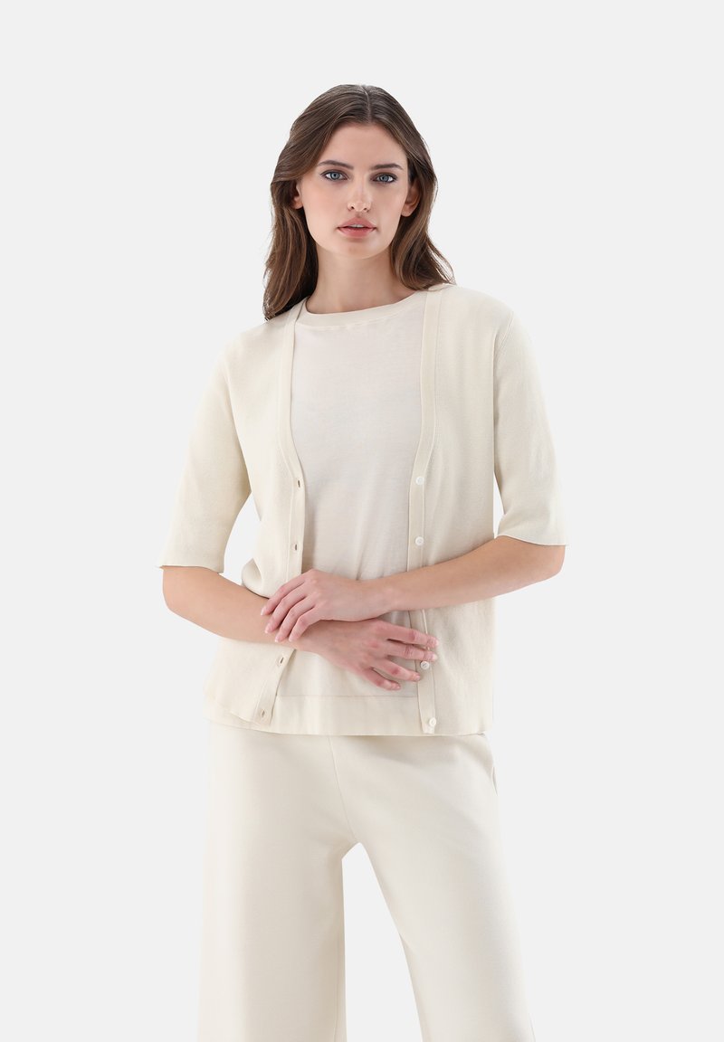 Helles beiges Cardigan mit kurzen Ärmeln, mit Knopfverschluss und weicher Textur, getragen über einem passenden cremefarbenen Oberteil und hellen Hosen.