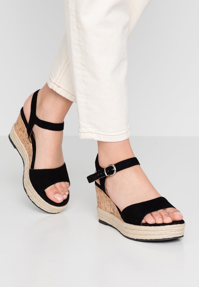 Pieds portant des sandales compensées en daim noir avec des brides à la cheville et un pantalon de couleur claire sur un fond uni.