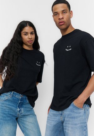 To unge voksne iført sorte T-shirts med teksten "NEJ TAK", kombineret med blå jeans, står foran en ensfarvet hvid baggrund.