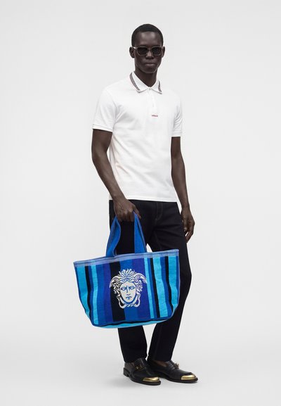 Homme portant des lunettes de soleil, un polo blanc, un pantalon noir et des chaussures noires tenant un grand sac cabas rayé bleu avec un motif de visage.