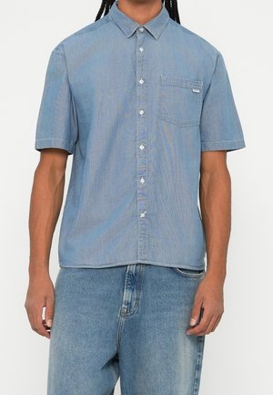 Mand iført lyseblå kortærmet skjorte med brystlomme kombineret med medium vasket blå jeans mod en hvid baggrund.