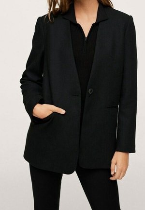Frau trägt einen schwarzen Blazer mit einem Knopf, eine Hand in der Tasche, über einem schwarzen Reißverschluss-Top und schwarzen Hosen.