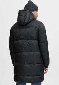 Abrigo acolchado negro con capucha, que presenta secciones acolchadas y puños elásticos. Textura suave, diseño hasta la rodilla y una silueta sencilla.