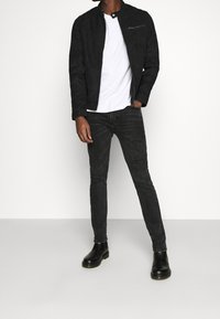 Chaqueta de ante negra con cuello alzado, bolsillo con cremallera; llevada sobre una camiseta blanca y combinada con jeans oscuros y botas negras hasta el tobillo.