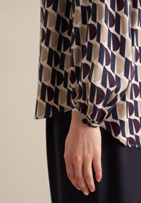 Blouse à manches longues en tissu crème avec un motif géométrique de formes marine, bordeaux et taupe. Détail de bouton au poignet.