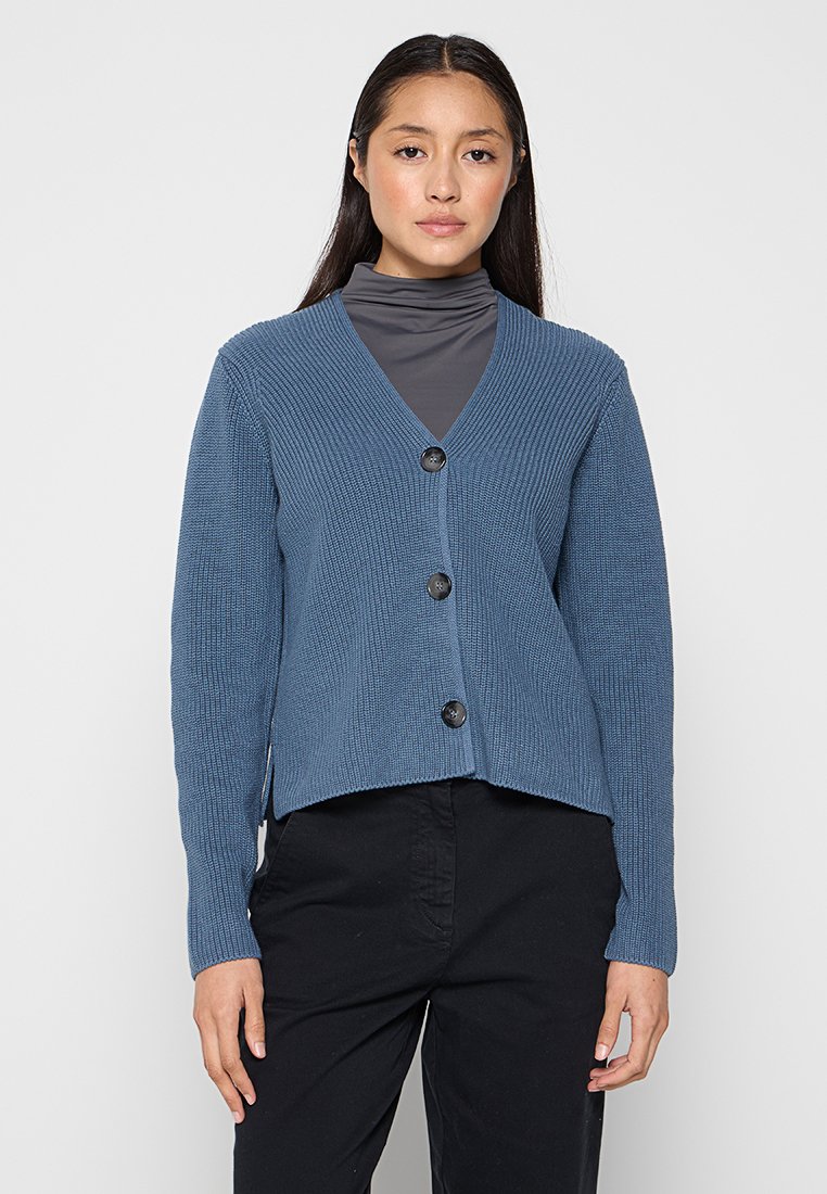 Blaue gerippte Strickjacke mit V-Ausschnitt, drei schwarzen Knöpfen und kurzes Design. Über einem grauen Rollkragenpullover getragen.