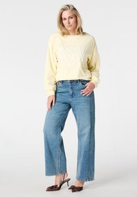 Geel sweatshirt met lange mouwen met de tekst "Positive Hearts", gecombineerd met lichtblauwe wijde broekspijpen met hoge taille en bruine pumps met stippen.