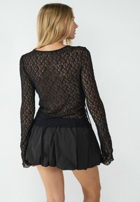 Schwarzes Spitzenshirt mit langen Ärmeln, das ein florales Muster und eine figurbetonte Passform aufweist, kombiniert mit einem schwarzen plissierten Rock für einen schichten Look.