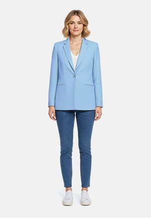 Vrouw die recht vooruit staat, gekleed in een lichtblauwe blazer, wit shirt, blauwe spijkerbroek en witte sneakers, op een effen witte achtergrond.