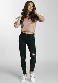 Lys rosa cropped genser, svarte skinny jeans med hull ved knærne, og hvite sneakers med gull detaljer. Jevn tekstur, avslappet passform.