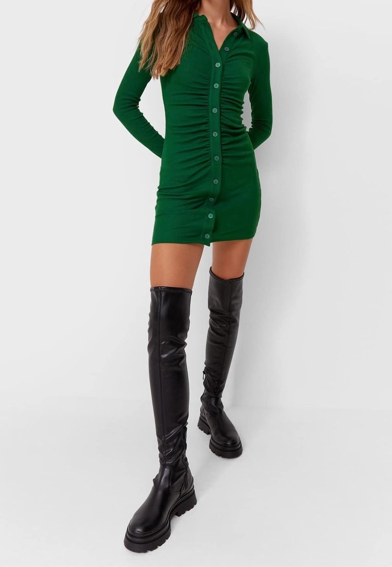 Femme portant une mini-robe verte cintrée à boutons avec fronces et des bottes en cuir épaisses noires au-dessus du genou, debout devant un fond uni.