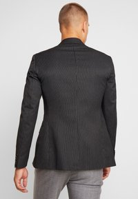 Blazer noir à rayures avec une coupe ajustée, doté d'un col cranté, d'un léger éclat et de deux fentes au dos. Porté sur un pantalon gris clair.