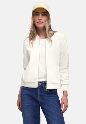 Kvinde iført en cremefarvet baseballkasket, cremefarvet lynlåstrøje, hvid skjorte og blå jeans, stående mod en ensartet lys baggrund.