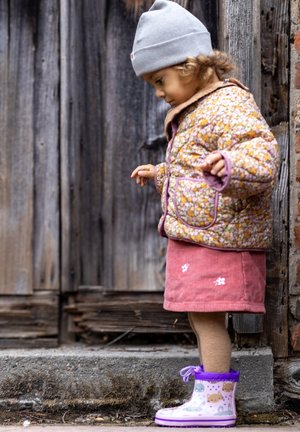 Gevulde bloemendruk jas in bruin en geel, roze corduroy rok met witte bloemenmotieven en paarse rubberen laarzen met dierenpatronen.