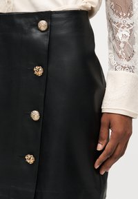 Μαύρη δερμάτινη φούστα με τέσσερα διακοσμητικά χρυσά κουμπιά, σε συνδυασμό με λευκό δαντελένιο μπλουζάκι με μακριά μανίκια και ένα σκουρόχρωμο χέρι που ακουμπά δίπλα της.