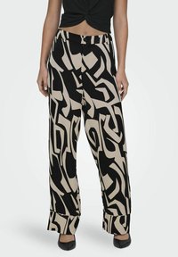 Pantalons à large jambes avec un motif abstrait noir et beige, portés avec un crop top noir torsadé et des chaussures à bout pointu noires sur fond uni.