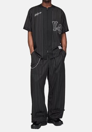 Uomo che indossa una camicia nera a righe sottili con zip e pantaloni abbinati a gamba larga, accessoriata con una catena e scarpe nere.