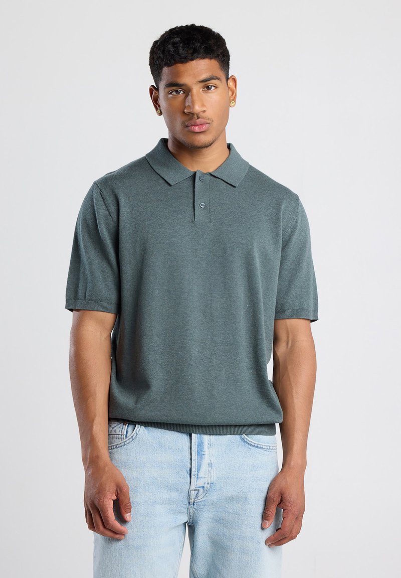 Polo de manga corta en verde apagado, hecho de una tela suave, presenta un collar clásico y una tapeta de tres botones en la parte delantera. Combinado con unos jeans de color azul claro.